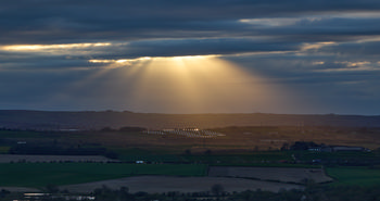 This landscape photograph captures a dramatic scene on a cloudy spring evening in the rural countryside of Derbyshire, United Kingdom, near Bolsover in England. The main subject is a solar farm, with rows of solar panels reflecting subtle light in the middle distance. Above the rolling fields, clouds dominate the sky, while striking beams of sunlight—often called Gods rays or crepuscular rays—break through the clouds, illuminating the scene below. The interplay of natural light with the modern solar farm highlights both the beauty of the English countryside and the presence of renewable energy in this rural area.